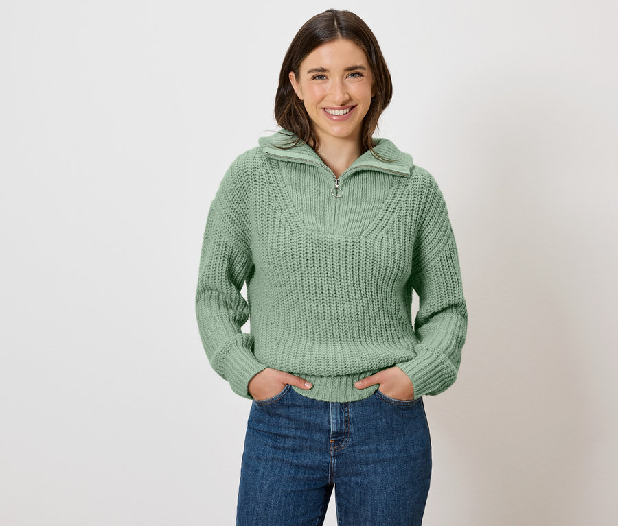 Frau mit braunen Haaren posiert in einem grünen Pullover und blauen Jeans.