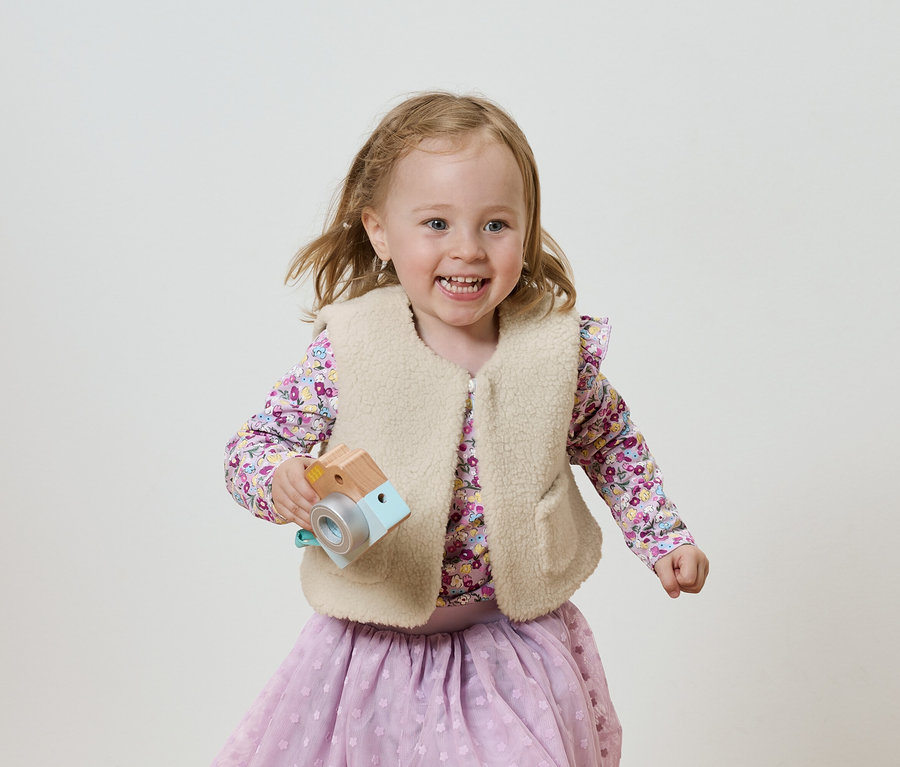 Lächelndes Mädchen mit Holzspielzeugkamera, gekleidet in eine fliederfarbene Bluse und einen Rock, vor einem hellen Hintergrund.