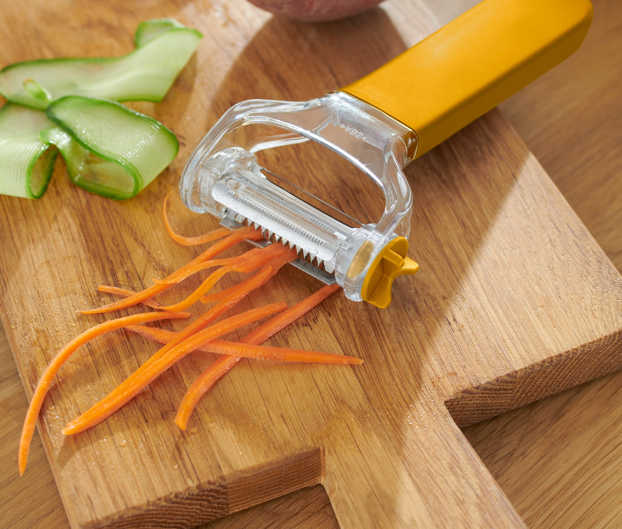 Gelber Gemüseschäler mit geschälter Karotte, Gurke und Kartoffel auf einem Holzbrett.
