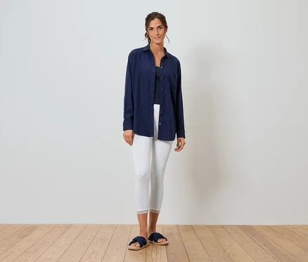 Frau steht vor einer weißen Wand und trägt ein blaues Hemd, weiße Leggings und blaue Hausschuhe.