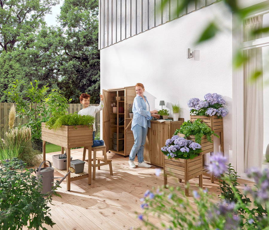 Ein Junge und eine Frau hantieren auf einer hölzernen Terrasse mit Gartenmöbeln und Pflanzen.