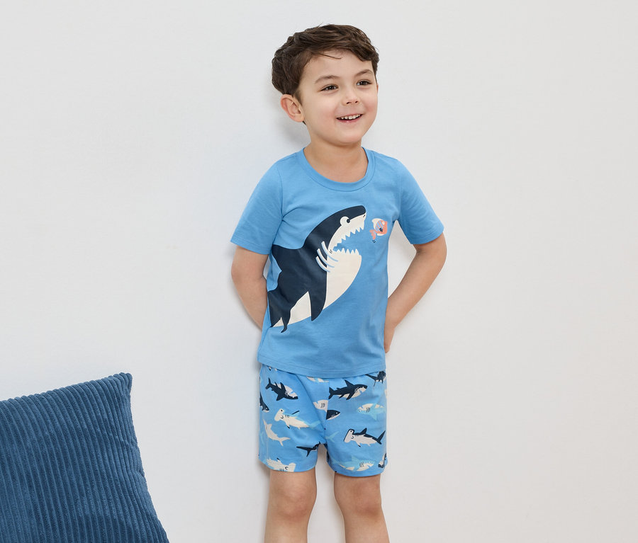 Ein lächelnder Junge trägt einen hellblauen Pyjama mit Hai-Motiv, vor einer weißen Wand.