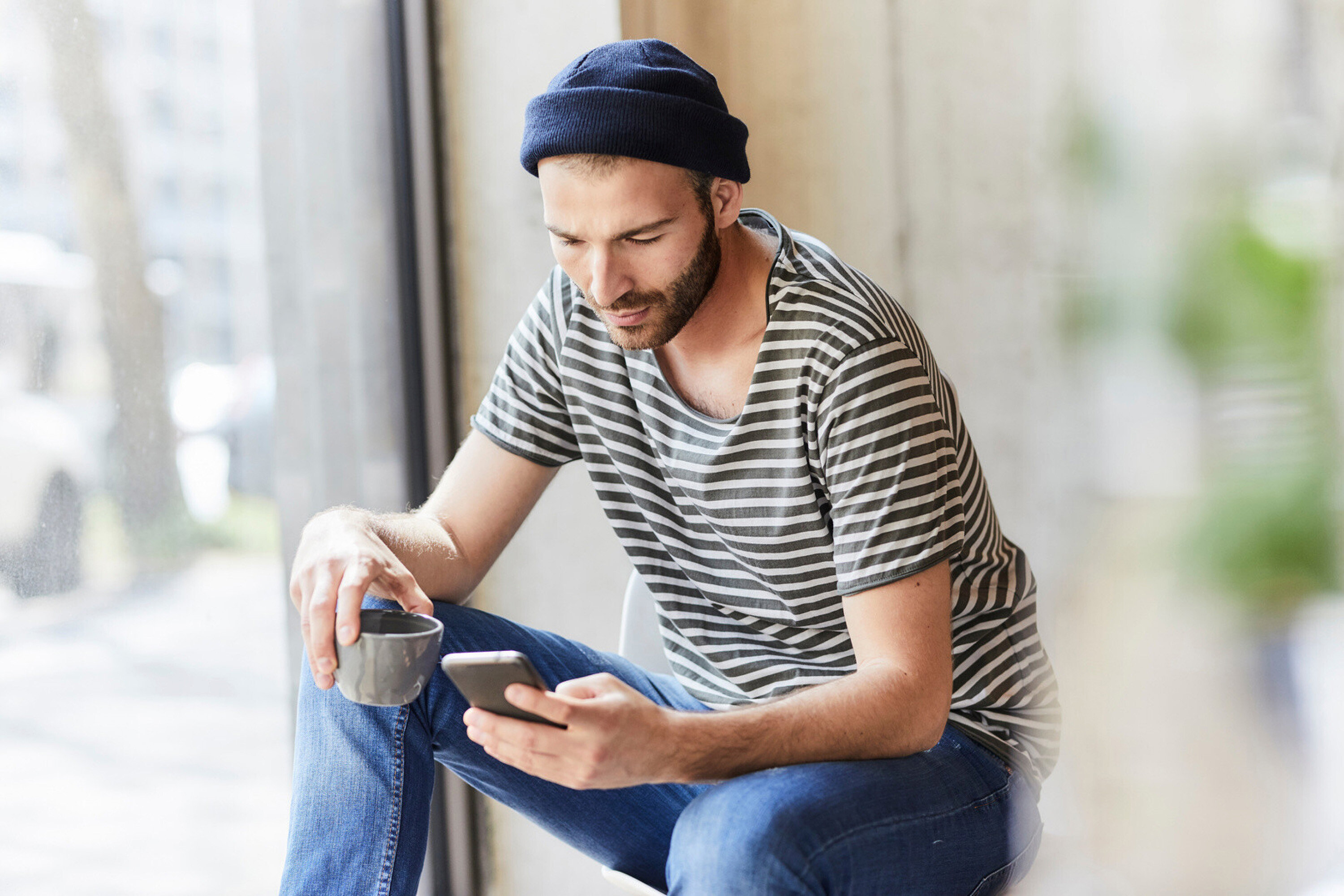 Mann mit Mütze und gestreiftem T-Shirt sitzt mit Handy und Tasse in der Hand.