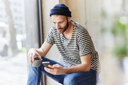 Mann mit Mütze und gestreiftem T-Shirt sitzt mit Handy und Tasse in der Hand.