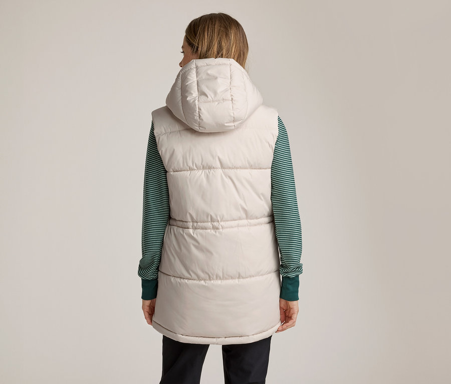 Eine Frau von hinten, die eine beigefarbene, wattierte Weste mit Kapuze über einem grün-weiß gestreiften Langarmshirt und dunklen Hosen trägt.