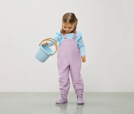 Mädchen mit hellblauer Bluse und lila Overall hält einen hellblauen Eimer.