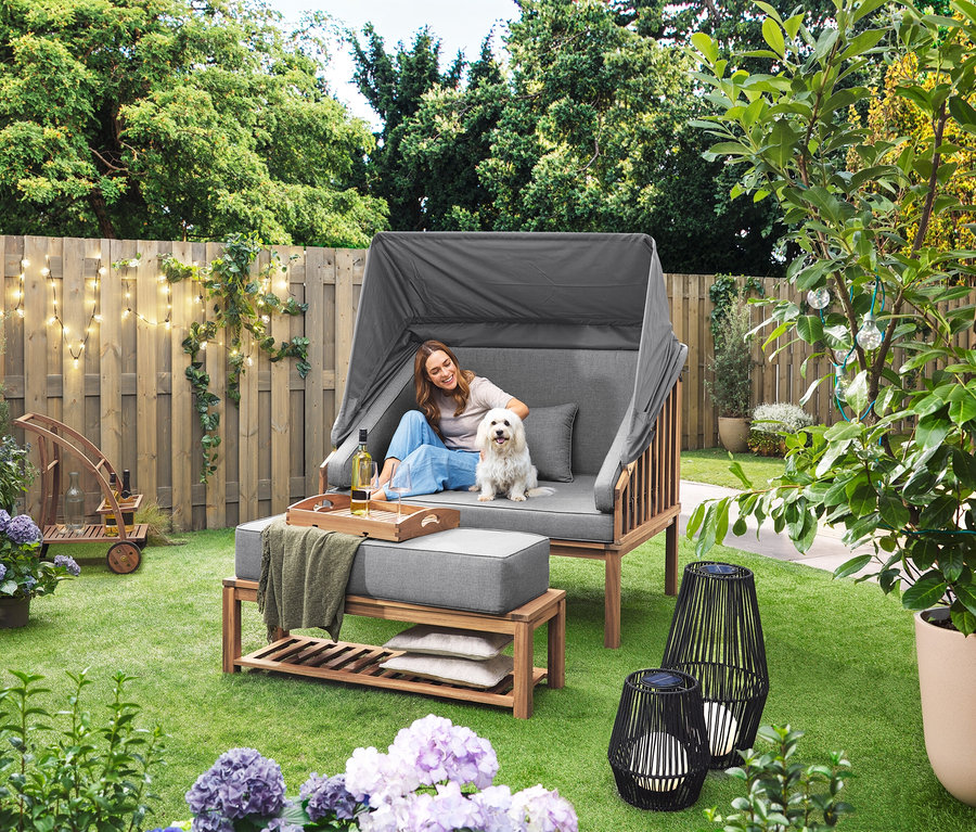 Eine Frau sitzt mit einem Hund in einem Premium Lounge-Strandkorb mit Sonnenschutz-Dach. Eine Solar-Laterne mit Textilband, groß und eine Solar-Laterne mit Textilband, klein stehen auf dem Rasen. Ein Barwagen mit abnehmbarem Tablett steht im Hintergrund.