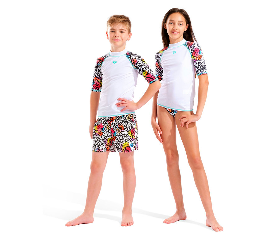 Zwei barfüßige Kinder, ein Junge und ein Mädchen, stehen nebeneinander vor einem weißen Hintergrund. Beide tragen weiße Oberteile mit farbigen Ärmeln und farbige Unterteile.