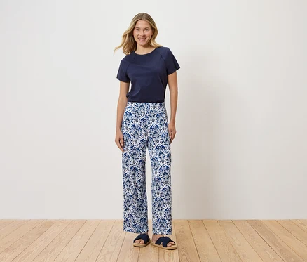 Eine Frau steht vor einer weißen Wand und trägt ein marineblaues T-Shirt, eine blaue und weiße Hose mit Blumenmuster und blaue Sandalen.