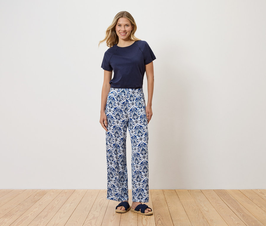 Eine Frau steht vor einer weißen Wand und trägt ein marineblaues T-Shirt, eine blaue und weiße Hose mit Blumenmuster und blaue Sandalen.