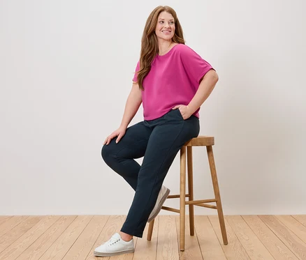 Frau sitzt auf einem Hocker, trägt ein pinkes Oberteil, eine blaue Hose und weiße Schuhe.