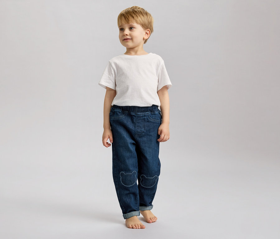 Ein Junge mit blonden Haaren steht barfuß in Jeans mit Bärenmotiven und einem weißen T-Shirt vor einem grauen Hintergrund.