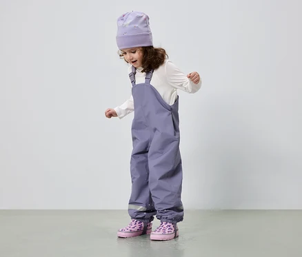 Ein kleines Mädchen mit lockigem Haar trägt eine lila Mütze, eine weiße Bluse, eine lila Hose und rosa Stiefel.