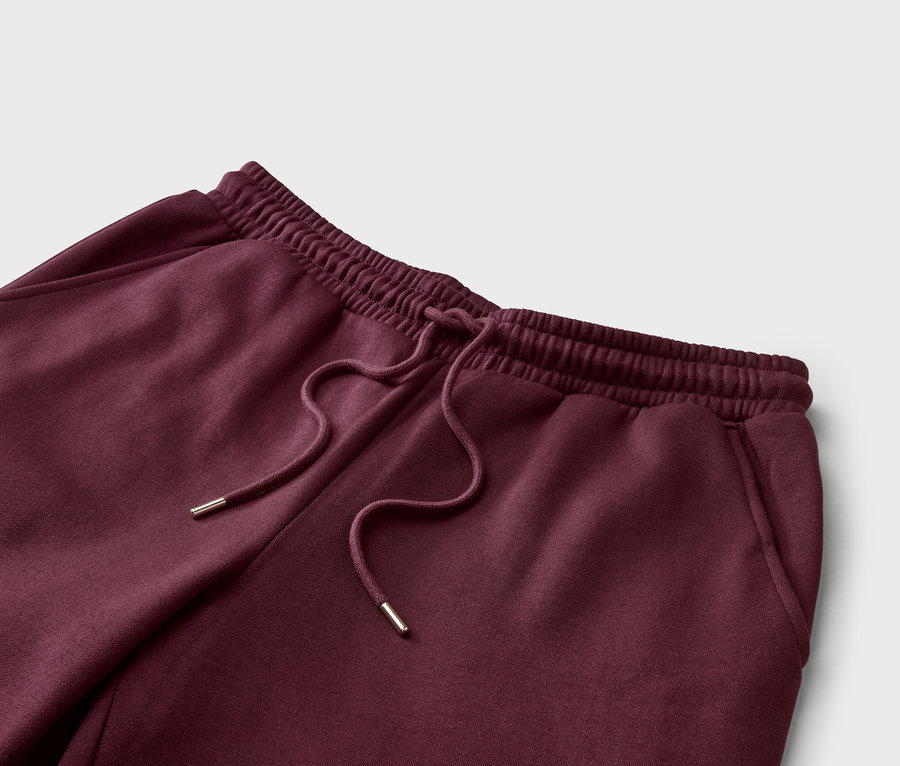 Nahaufnahme einer bordeauxroten Jogginghose mit Kordelzug und elastischem Bund vor einem hellgrauen Hintergrund.