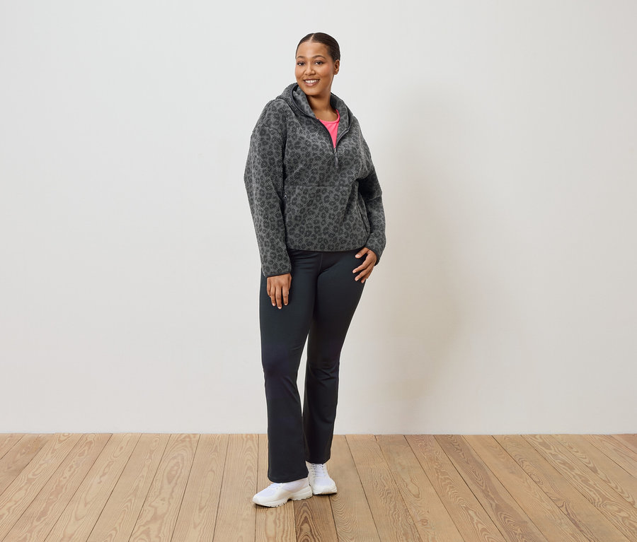 Frau steht und trägt einen grau-schwarzen Leopard-Fleece-Hoodie, schwarze Hosen und weiße Turnschuhe.