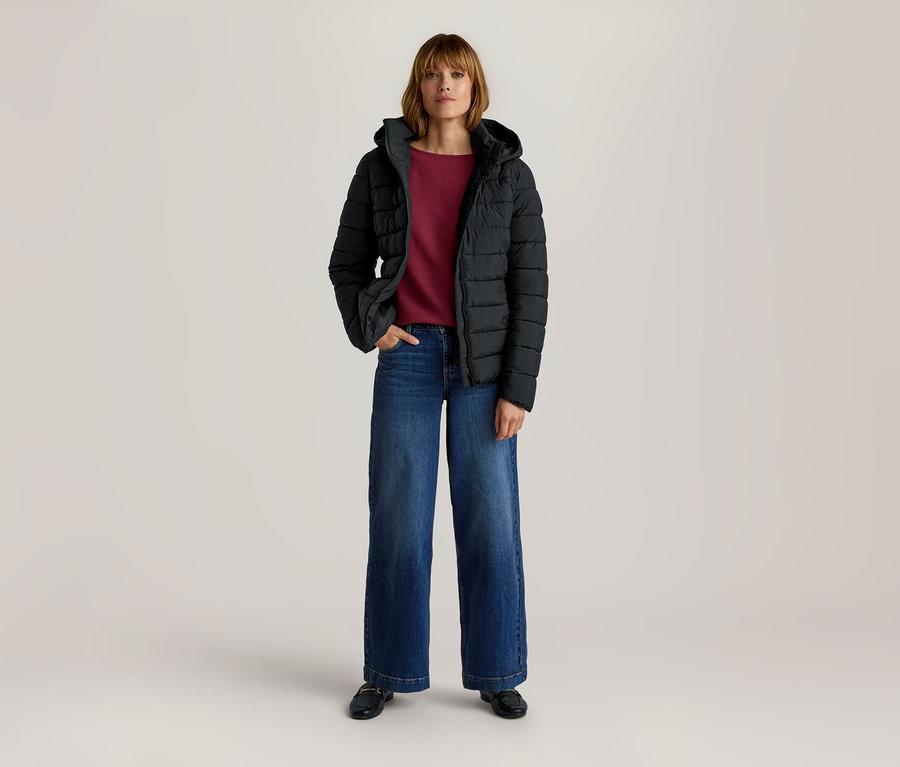 Frau steht in schwarzer Kapuzen-Steppjacke, rotem Langarmshirt, blauer weiter Jeans und schwarzen Slippern, eine Hand in der Tasche.