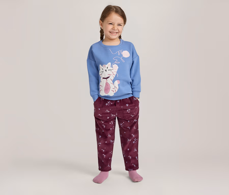 Lächelndes Mädchen mit Zöpfen, das ein blaues Langarmshirt mit Katzenmotiv und eine weinrote Hose mit Blumenmuster trägt.