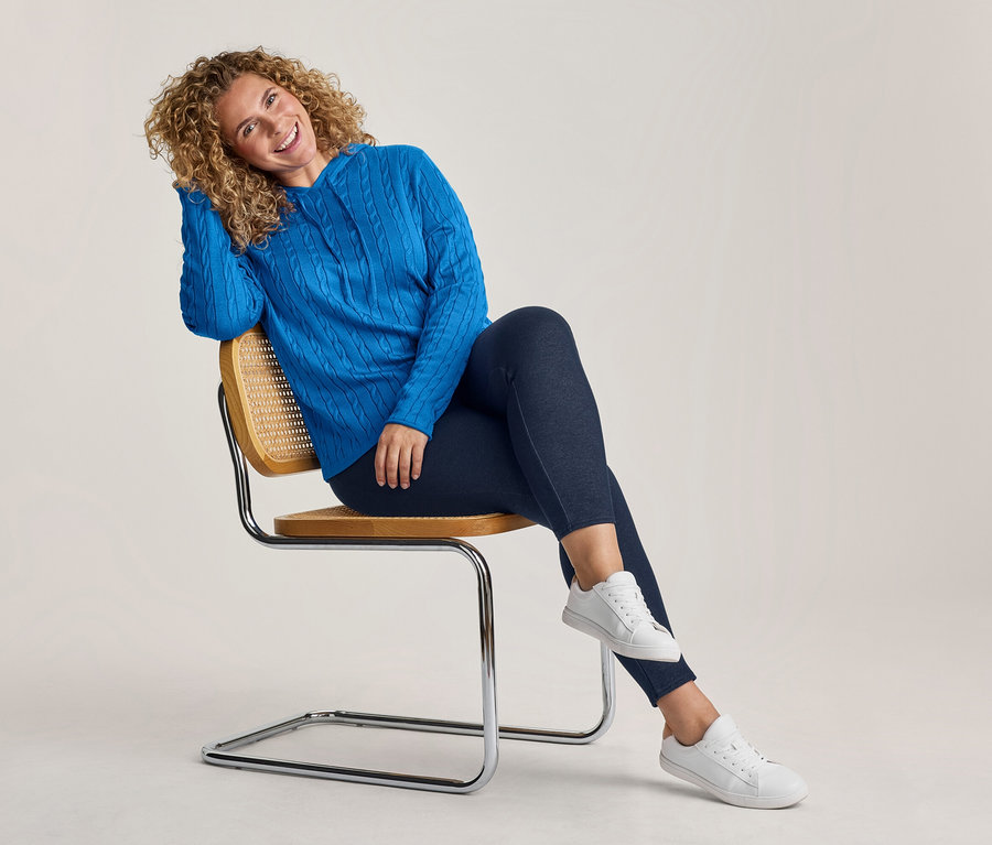 Lächelnde Frau mit lockigem Haar sitzt auf einem Stuhl, trägt einen blauen Zopfstrick-Kapuzenpullover, dunkle Hose und weiße Sneaker.
