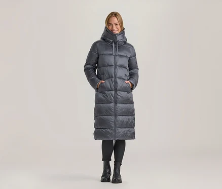 Lächelnde Frau steht in einem langen grauen Steppmantel mit den Händen in den Taschen, schwarzen Leggings und schwarzen Stiefeln.