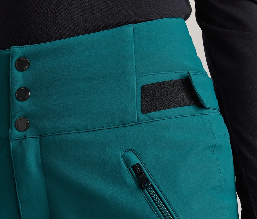 Nahaufnahme einer Person in hochgeschnittenen, türkisfarbenen Hosen und einem schwarzen Oberteil. Die Hose hat drei schwarze Knöpfe, eine Reißverschlusstasche und eine Patte mit Klettverschluss.