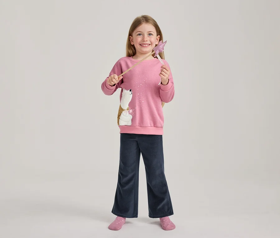 Lächelndes Mädchen mit langen Haaren trägt ein pinkes Sweatshirt mit einem Pailletten-Eisbären, dunkelblaue weite Hosen und pinke Socken, hält einen pinken Sternenzauberstab.