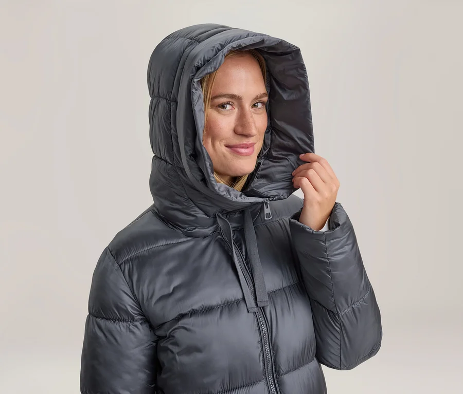 Lächelnde Frau in einer dunkelgrauen Steppjacke mit Kapuze, die sie mit einer Hand festhält.