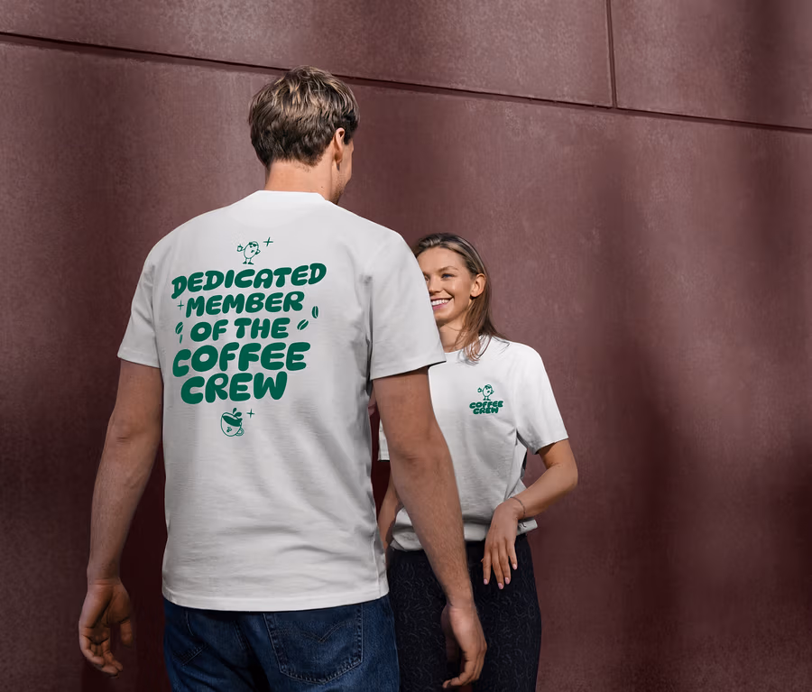Ein Mann trägt ein weißes Unisex Coffee Crew T-Shirt mit grünem Aufdruck "DEDICATED MEMBER OF THE COFFEE CREW" auf dem Rücken. Eine Frau lächelt im Hintergrund und trägt ebenfalls ein weißes T-Shirt mit dem "Coffee Crew"-Logo.