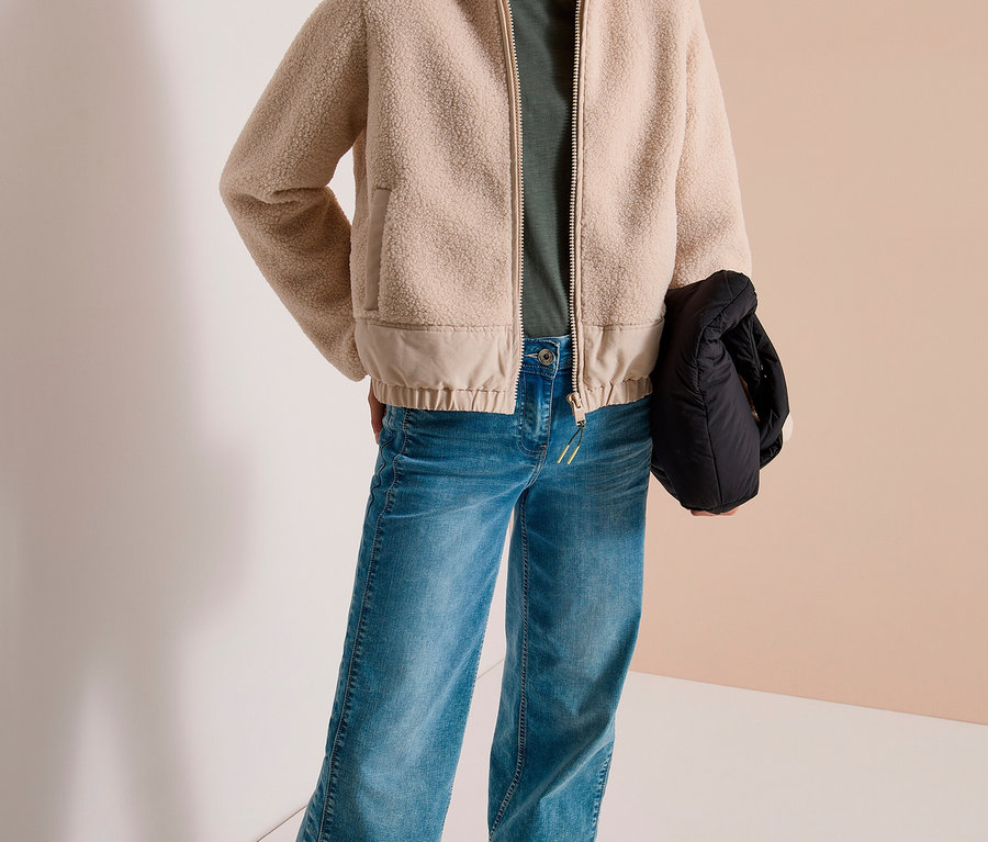 Eine lächelnde Frau steht in einer beigefarbenen CECIL Jacke mit Kapuze, grünem T-Shirt, blauen Jeans und weißen Sneakern. Sie hält eine schwarze Tasche.