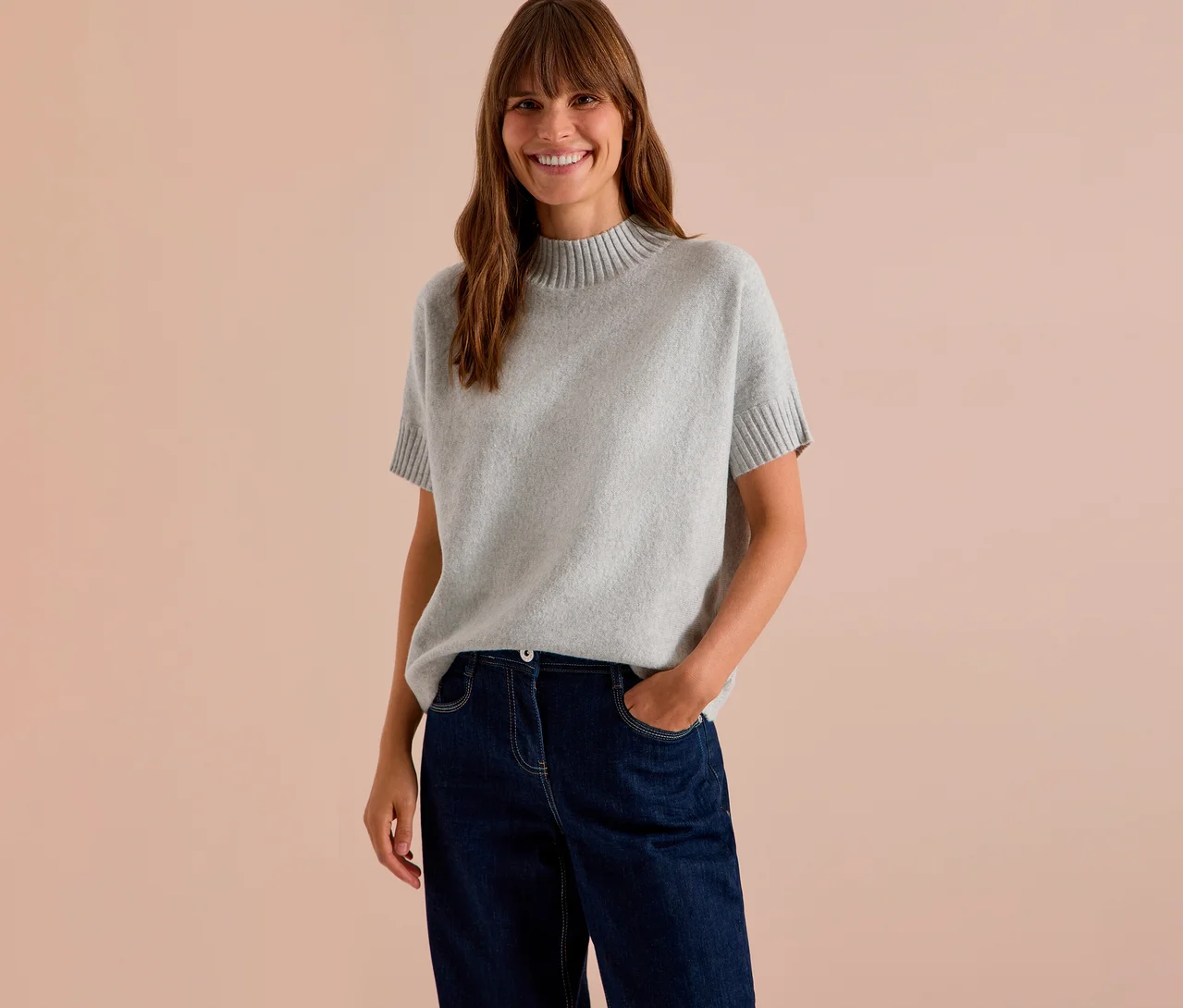 Lächelnde Frau mit langen braunen Haaren trägt einen grauen CECIL Strickpullover mit kurzen Ärmeln und eine dunkle Jeans.
