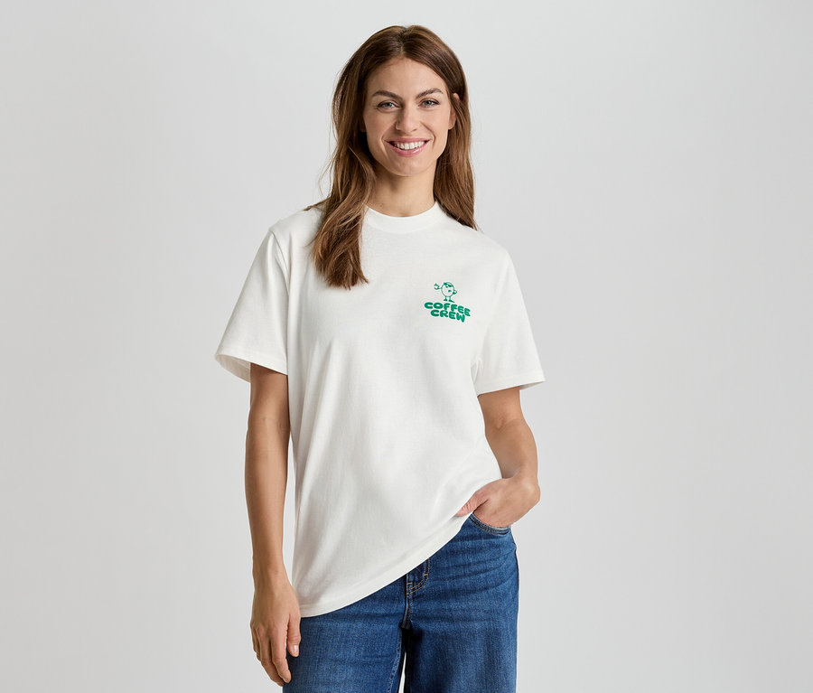 Frau mit weißem T-Shirt mit "Coffee Crew" Aufdruck und blauen Jeans.