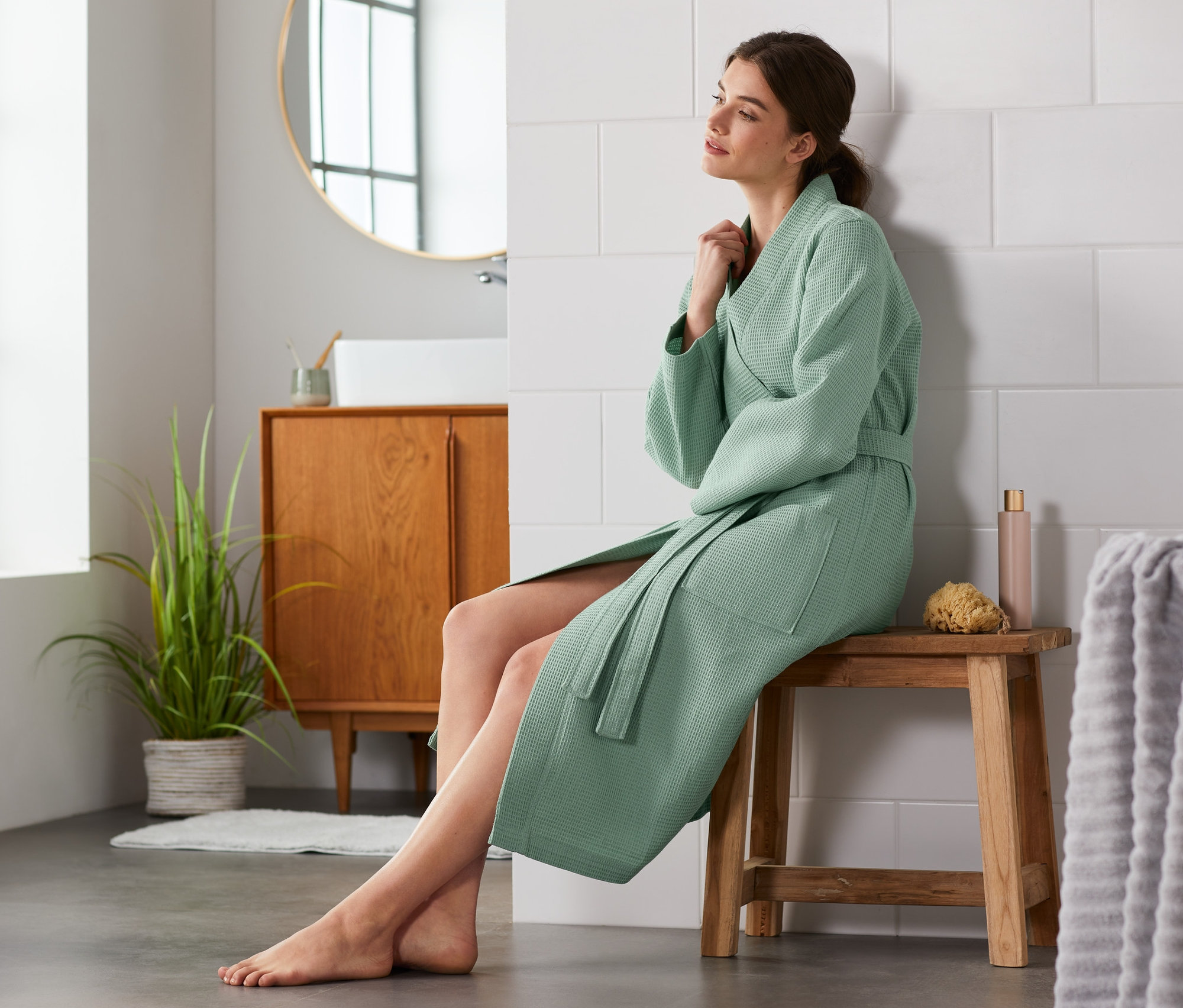 Frau sitzt im Damen-Bademantel in Waffelpiqué auf einem Hocker im Badezimmer.