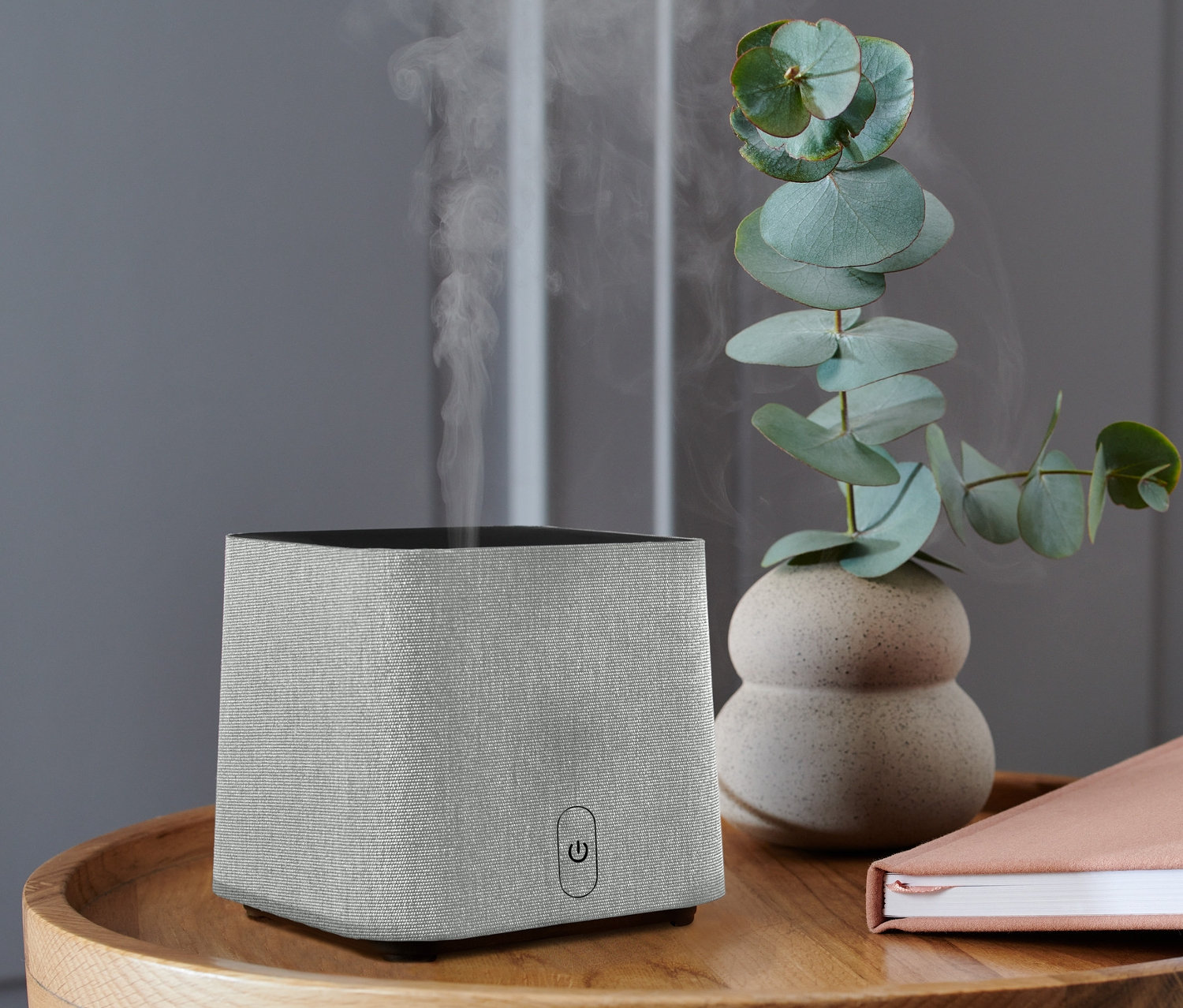 Ein grauer Aromadiffusor steht auf einem Holztisch, zusammen mit einem Buch und einer Eukalyptusvase.