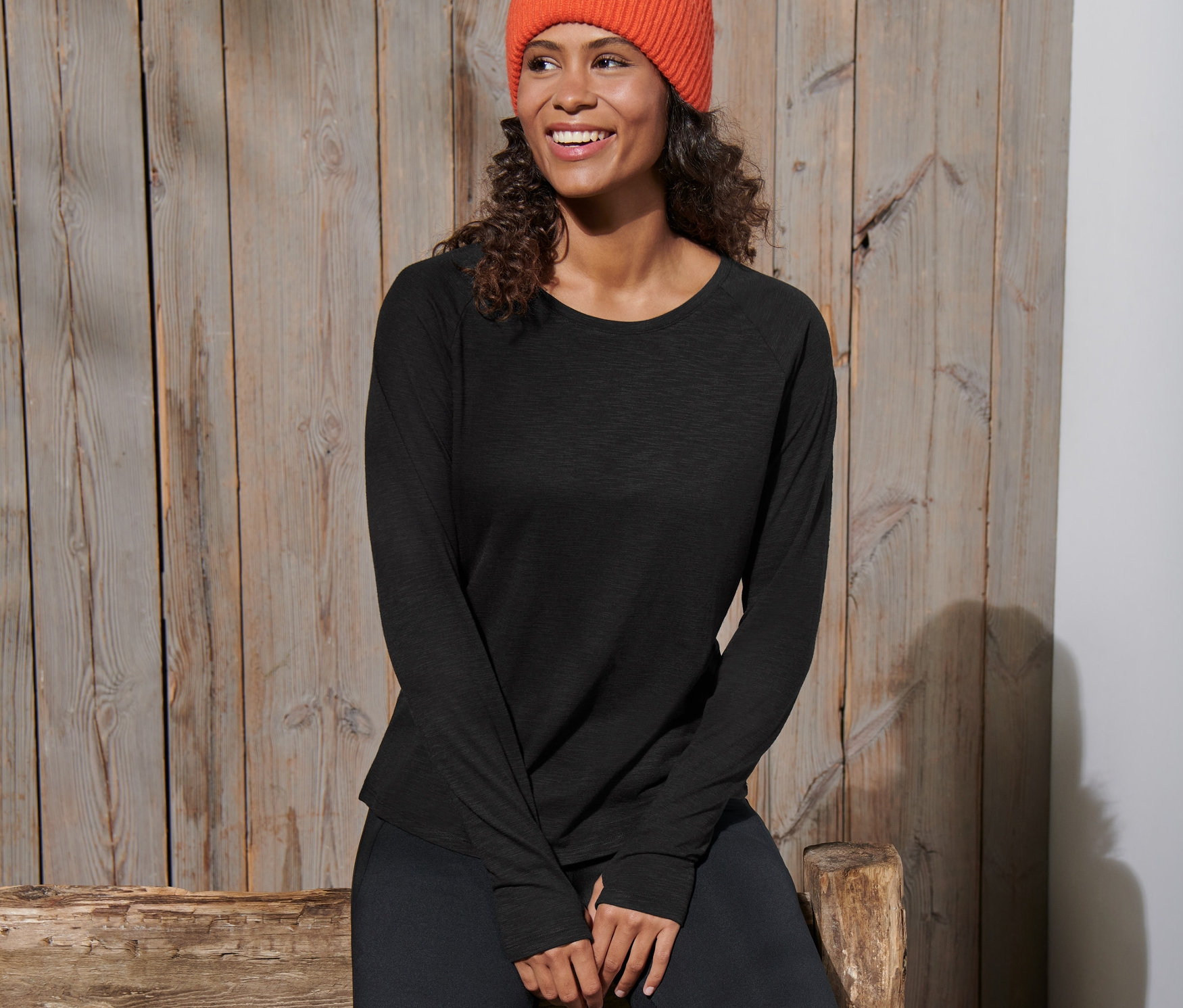 Frau sitzt auf einer Holzbank und trägt ein schwarzes Langarm-Sportshirt und eine orangefarbene Mütze.