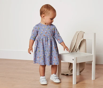 Ein Baby steht und trägt ein Baby-Jerseykleid mit Blumenmuster.