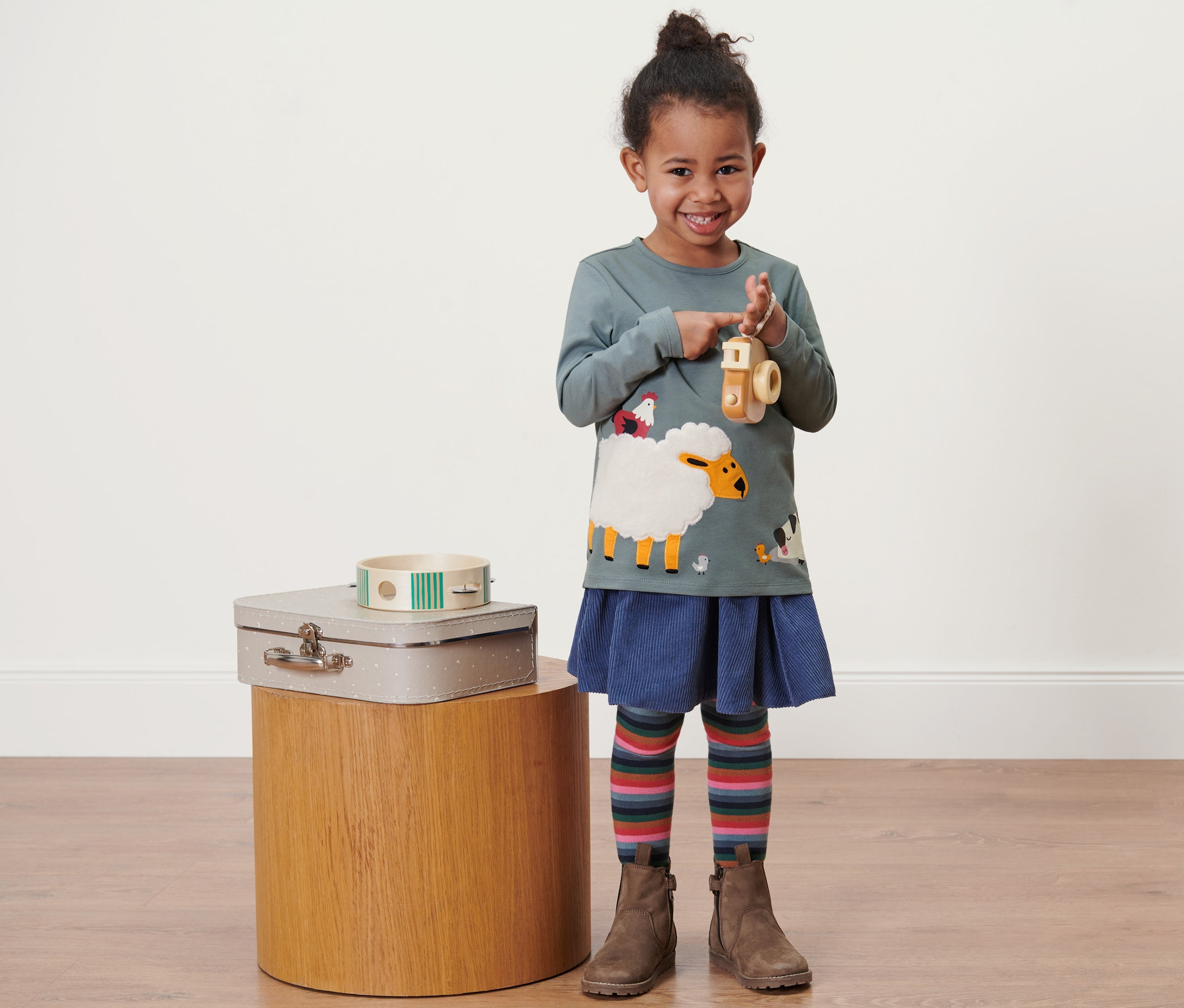 Ein lächelndes kleines Mädchen hält eine Holzkamera und trägt einen blauen Rock, bunte Leggings und braune Stiefel. Sie trägt 2 Kleinkind-Langarmshirts mit einem Schafmotiv.