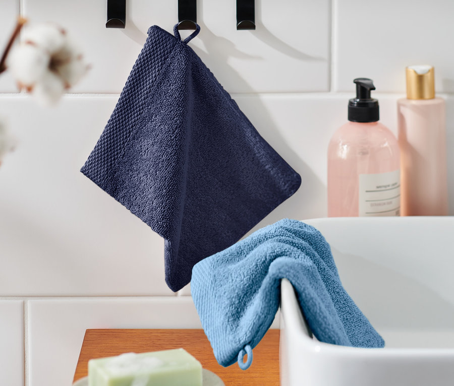 Ein dunkelblauer Waschlappen hängt an einem Haken neben einem hellblauen Waschlappen auf einem Waschbecken. Im Hintergrund Kosmetikflaschen und Blumen. 2 Waschlappen.