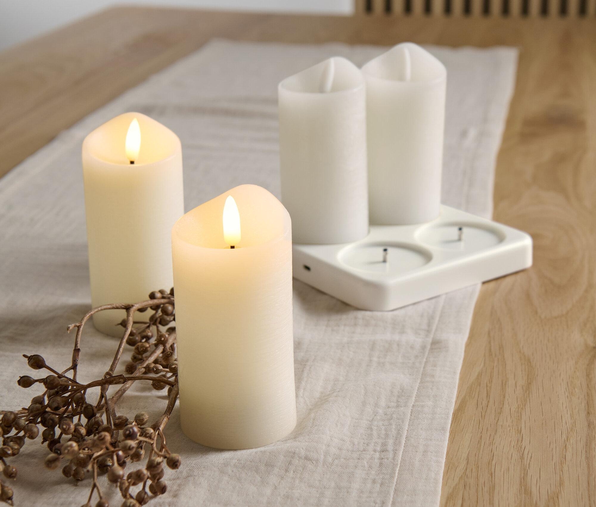 Auf einem Tisch liegen 4 LED-Akku-Echtwachskerzen mit Ladestation und ein Zweig mit Beeren.