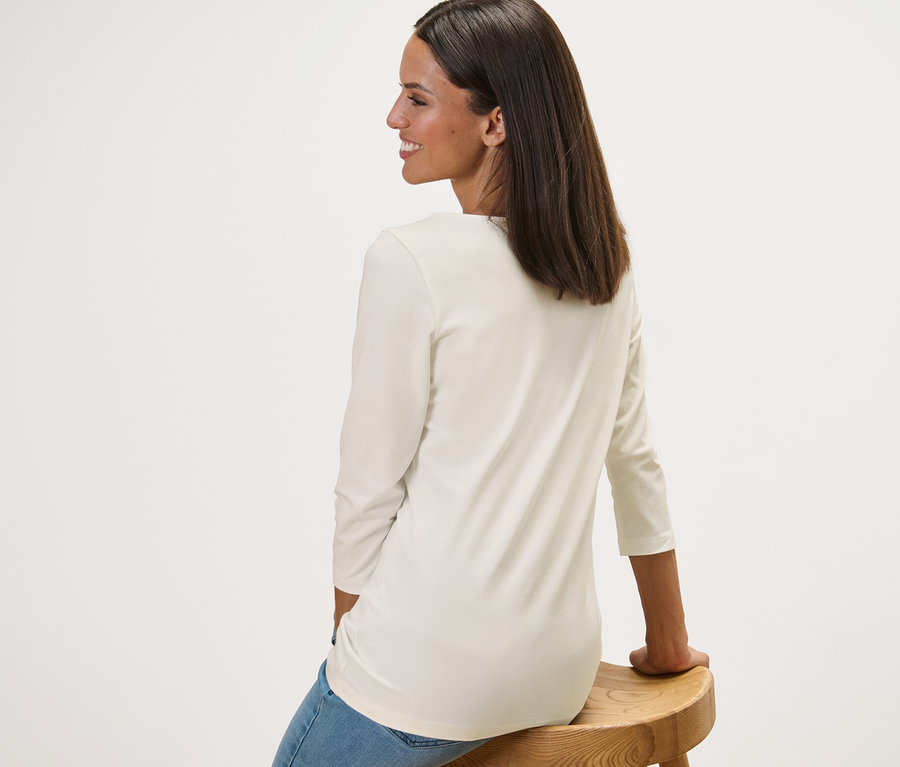 Eine Frau sitzt seitlich auf einem Holzschemel und trägt ein weißes Shirt mit 3/4-Arm.
