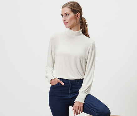Frau posiert mit einem Bein auf einem Holzhocker, trägt ein cremefarbenes Blusenshirt mit Stehkragen und Jeans.