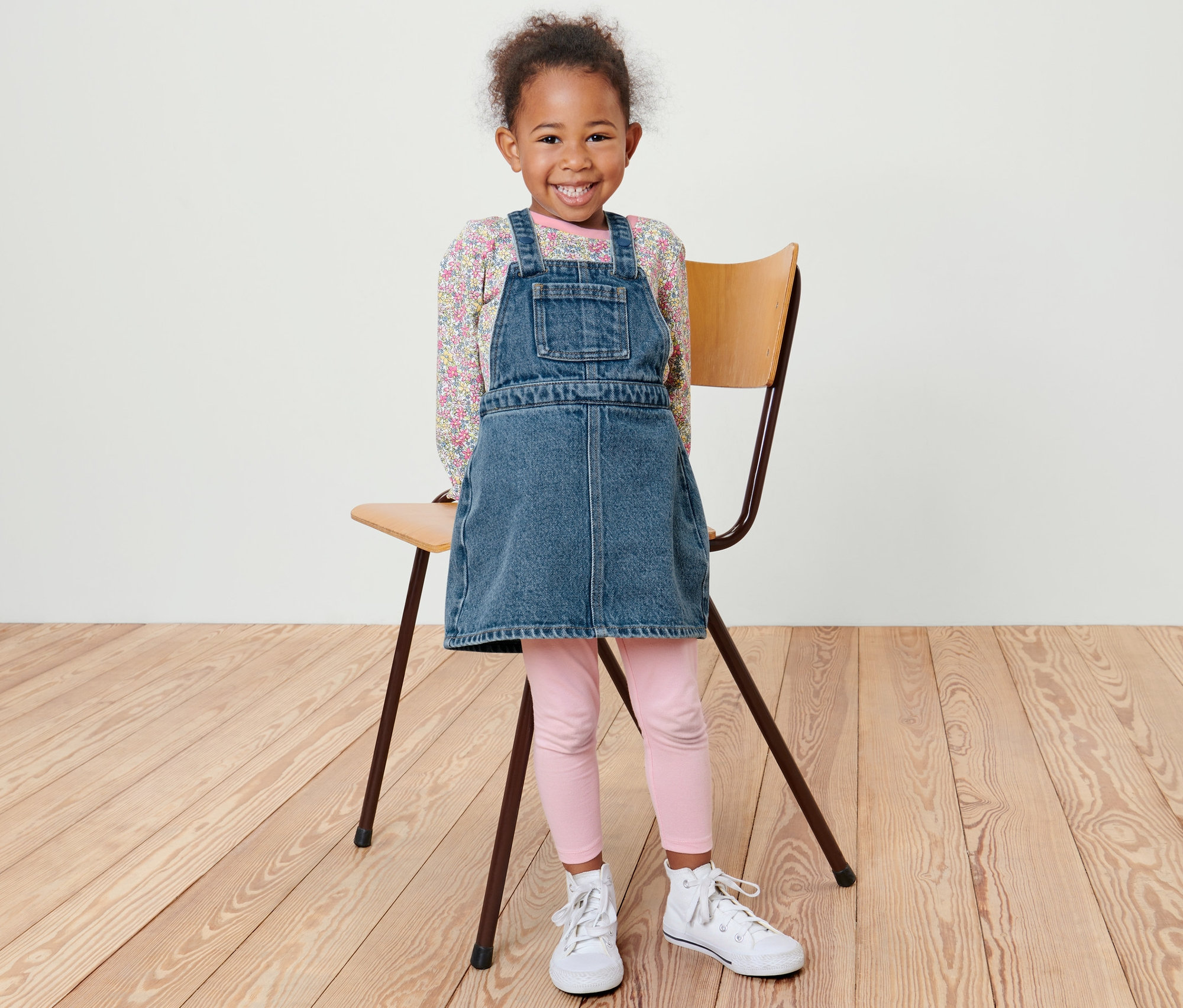 Ein lächelndes kleines Mädchen steht neben einem Stuhl und trägt ein Kleinkind-Latzkleid, eine rosa Leggings und weiße Turnschuhe.