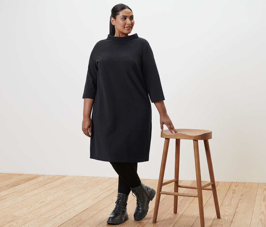 Frau in schwarzen Leder-Boots und Sweatkleid mit 3/4-Arm, die neben einem Holzhocker steht.