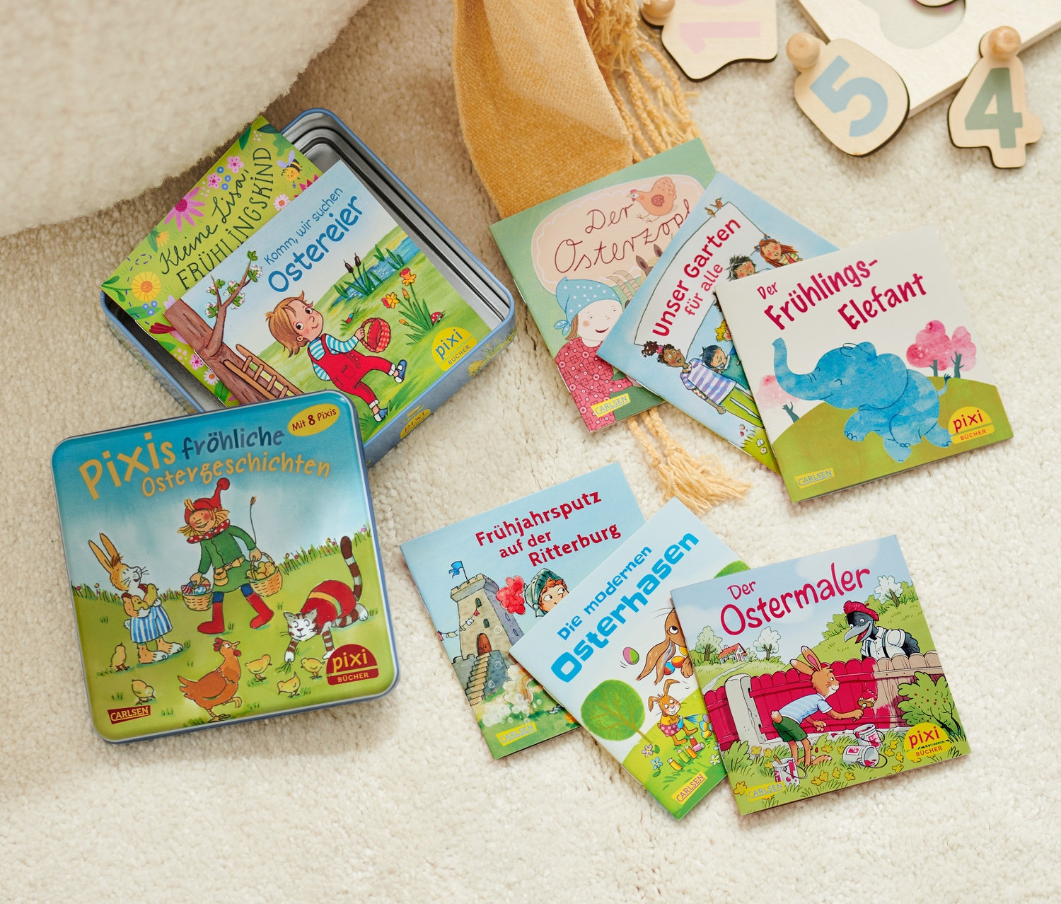 Eine Metalldose mit Pixi-Büchern und ein hölzernes Zahlenpuzzle liegen auf einem zotteligen Teppich.