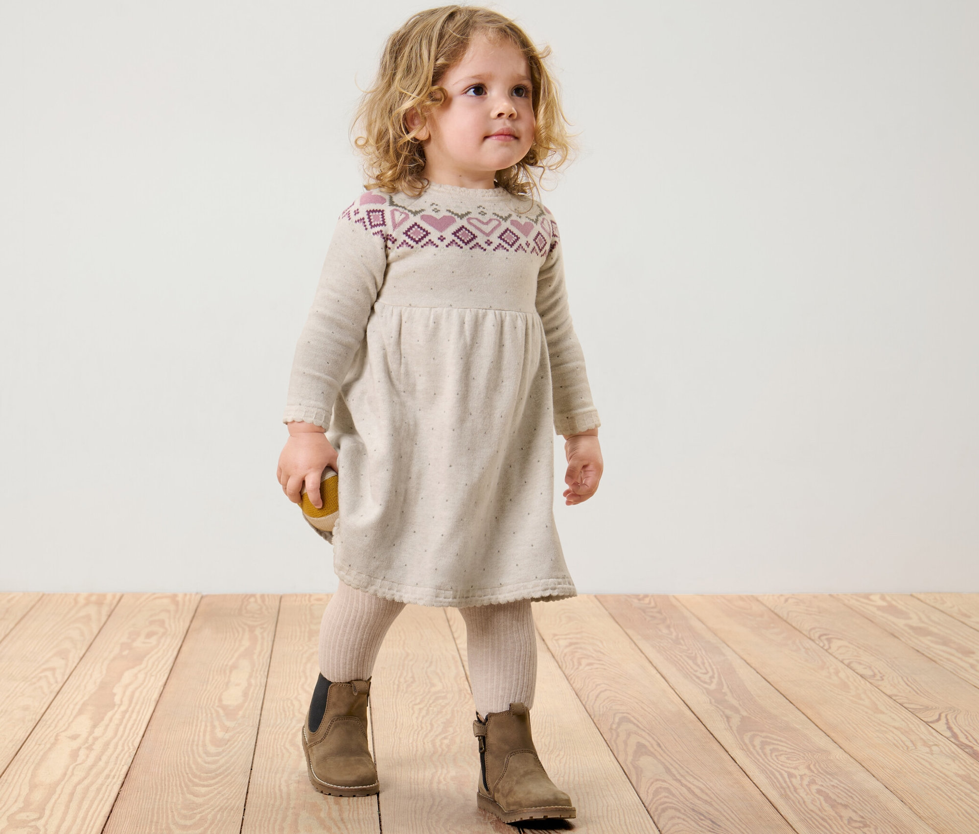 Ein Mädchen steht auf einem Holzboden und trägt ein Kinder-Strick-Kleid.