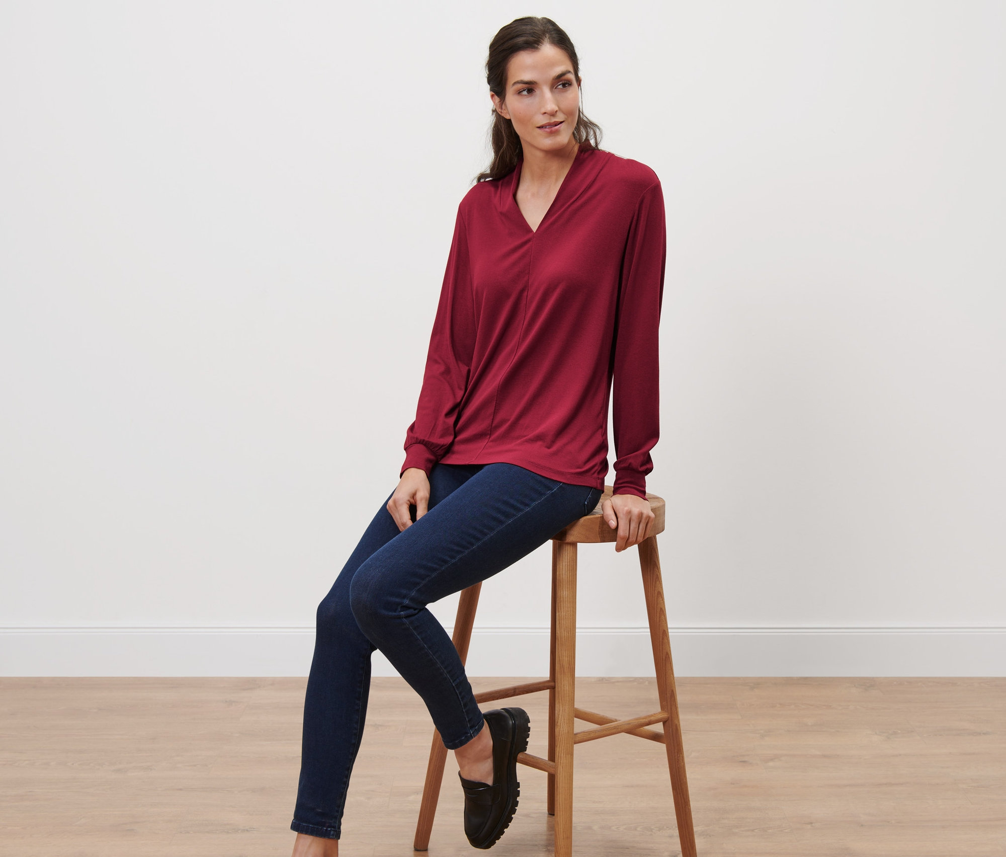 Frau sitzt auf einem Hocker und trägt ein bordeauxrotes Langarmshirt mit V-Ausschnitt.