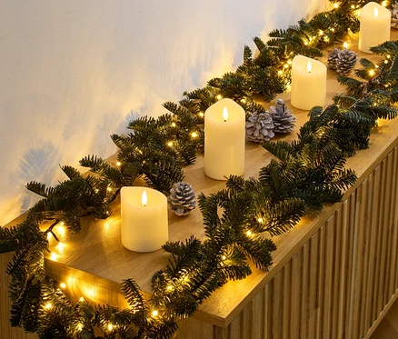 LED-Tannengirlande mit Kerzen und Tannenzapfen liegt auf einer Holzkommode.