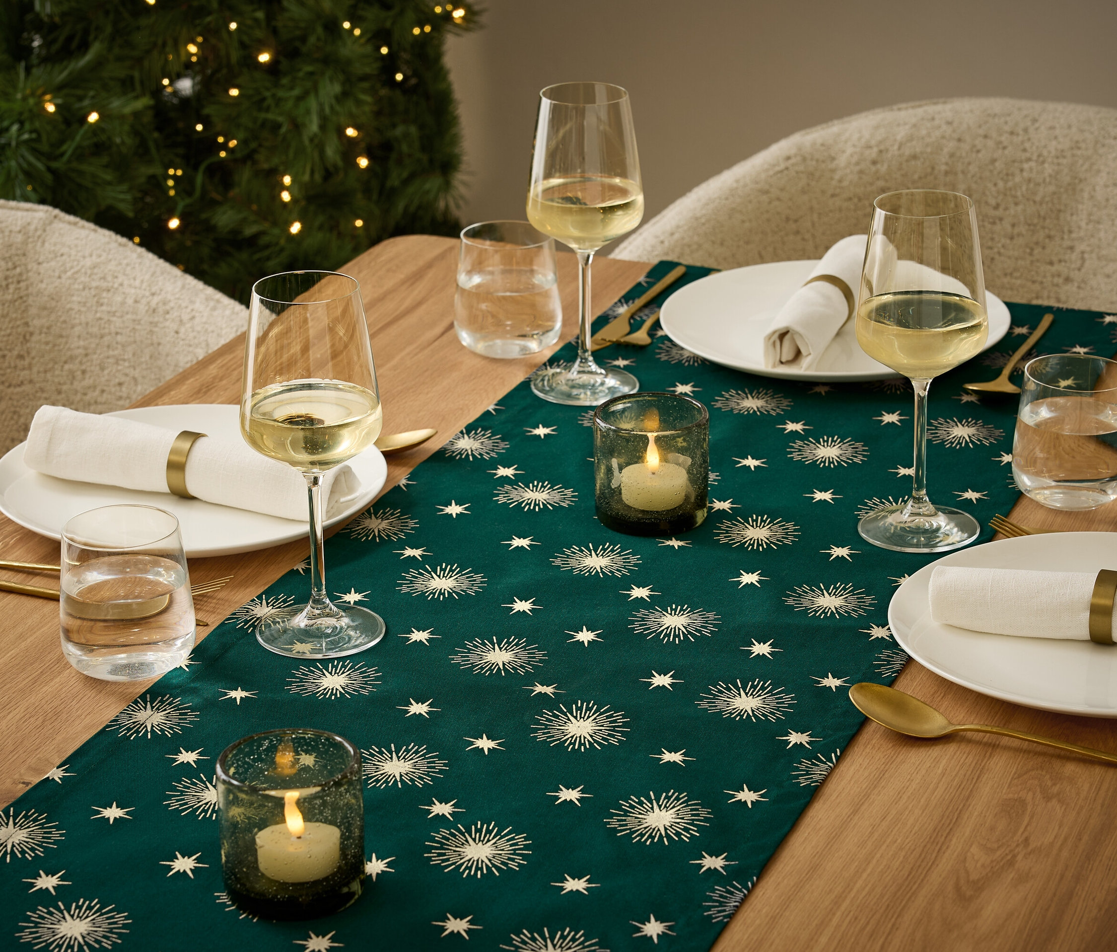 Festlich gedeckter Tisch mit einem dunkelgrünen Läufer mit Sternenmuster, Tellern mit Servietten, Gläsern mit Wein und Wasser und Kerzenhaltern mit brennenden Kerzen.