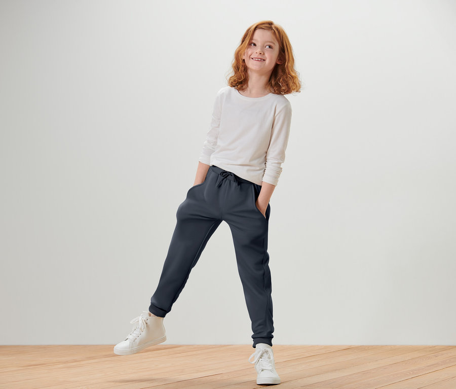 Rothaariges Mädchen in einem weißen Langarmshirt und dunkelblauer Jogginghose steht auf einem Bein und lächelt.