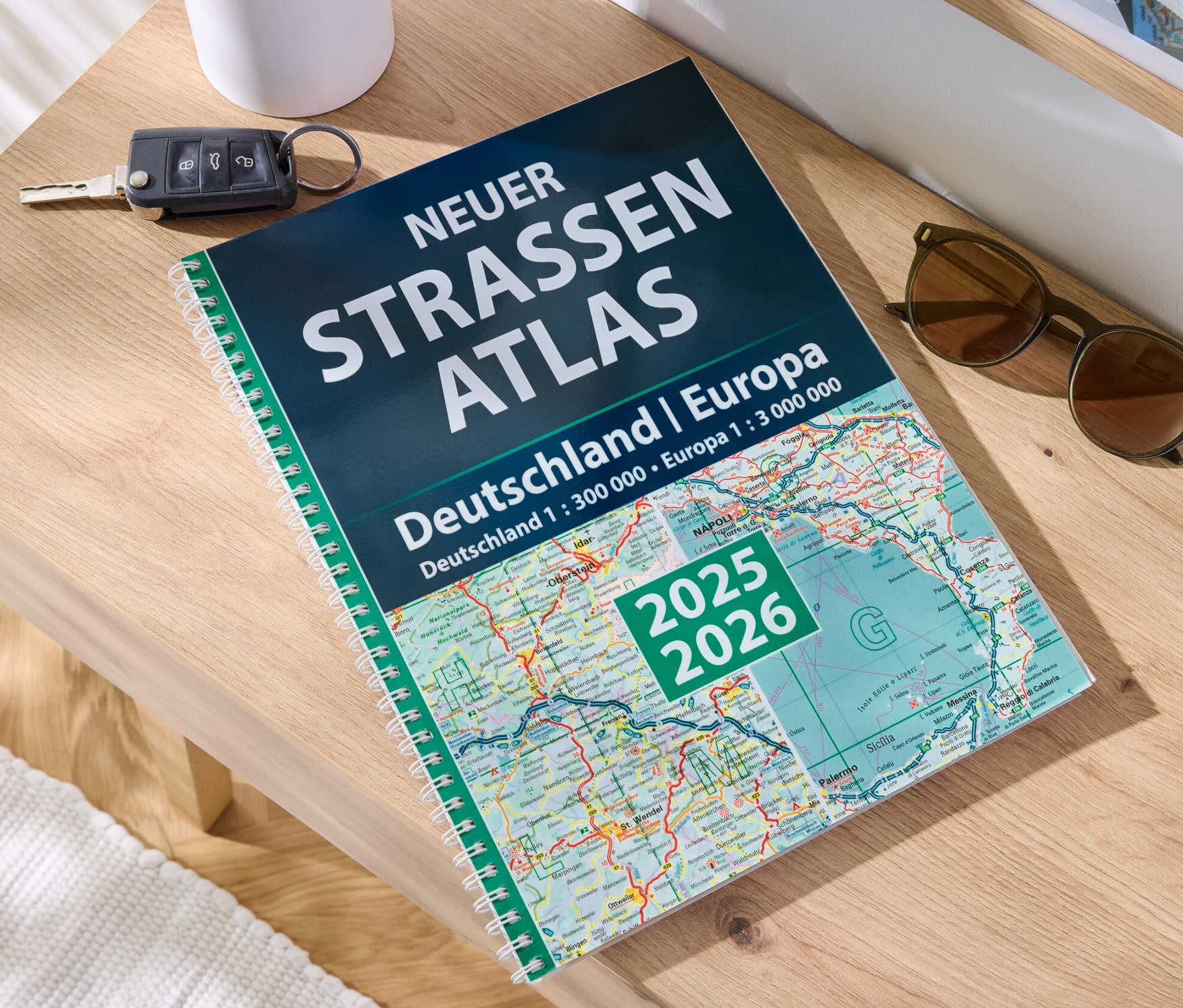 Auf einem Holztisch liegen ein Buch 'Neuer Straßen-Atlas Deutschland/Europa 2025/2026', ein Autoschlüssel und eine Sonnenbrille.