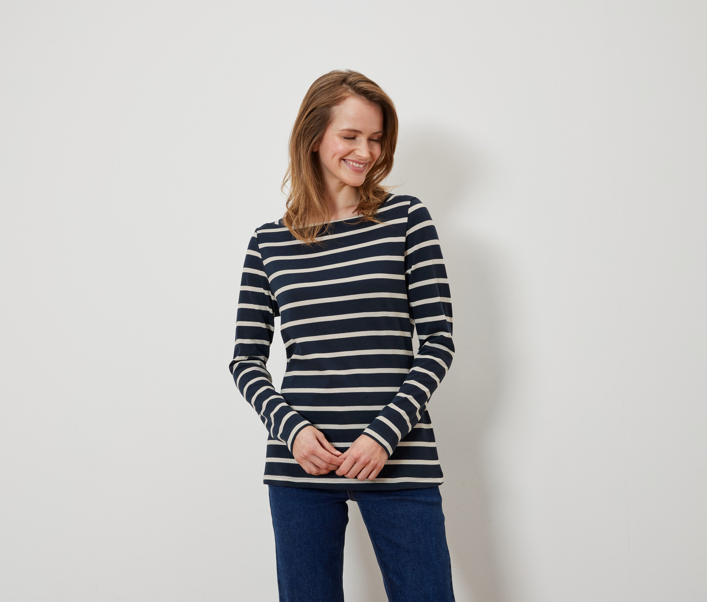 Eine Frau mit braunen Haaren und blauen Augen trägt ein blau-weiß gestreiftes Langarmshirt und blaue Jeans.