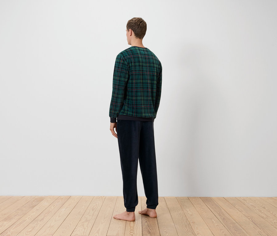 Ein Mann mit dem Rücken zur Kamera trägt ein grün-schwarz kariertes Oberteil und dunkelblaue Hosen und steht auf einem Holzboden vor einer weißen Wand.